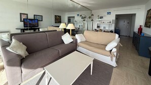 A living room with a brown sofa, a beige sofa, and a white coffee table. There is a TV on a stand, a lamp, and a bookshelf. - 06DJ - Apartment with sea view for 4 people - terrace (Villeneuve-Loubet)