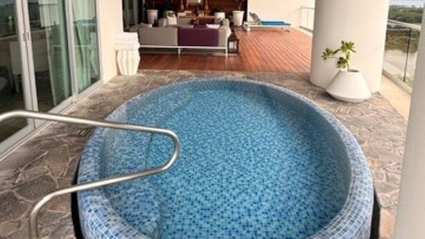 Una piscina con bordo piastrellato, circondata da pavimento in pietra, e vista su un interno moderno con divano e zona pranzo.