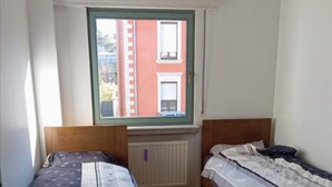 Una stanza piccola con due letti singoli, una finestra con vista sugli edifici e un termosifone.