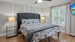 Ein Schlafzimmer mit einer gepolsterten Kopfteile, einem Bett mit gemusterten Bettbezügen, zwei Nachttischlampen, einem Deckenventilator und einem Fenster mit Vorhängen.