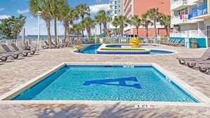 Ein Swimmingpool mit dem Buchstaben 'A' im Wasser, umgeben von Liegestühlen und Palmen, mit einem Hochhaus im Hintergrund.