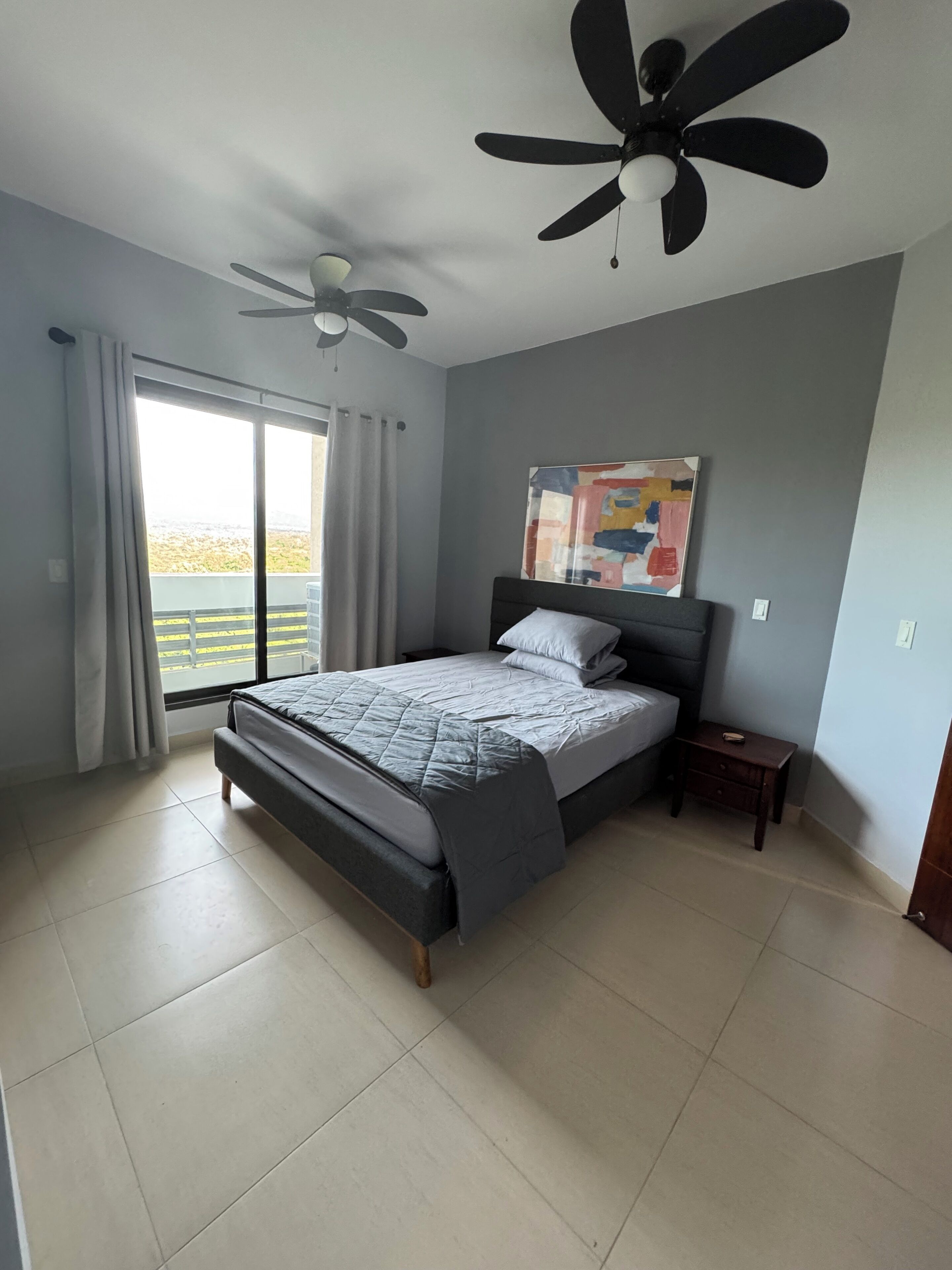 Un dormitorio con cama, ventilador de techo, mesita de noche y ventana con cortinas.