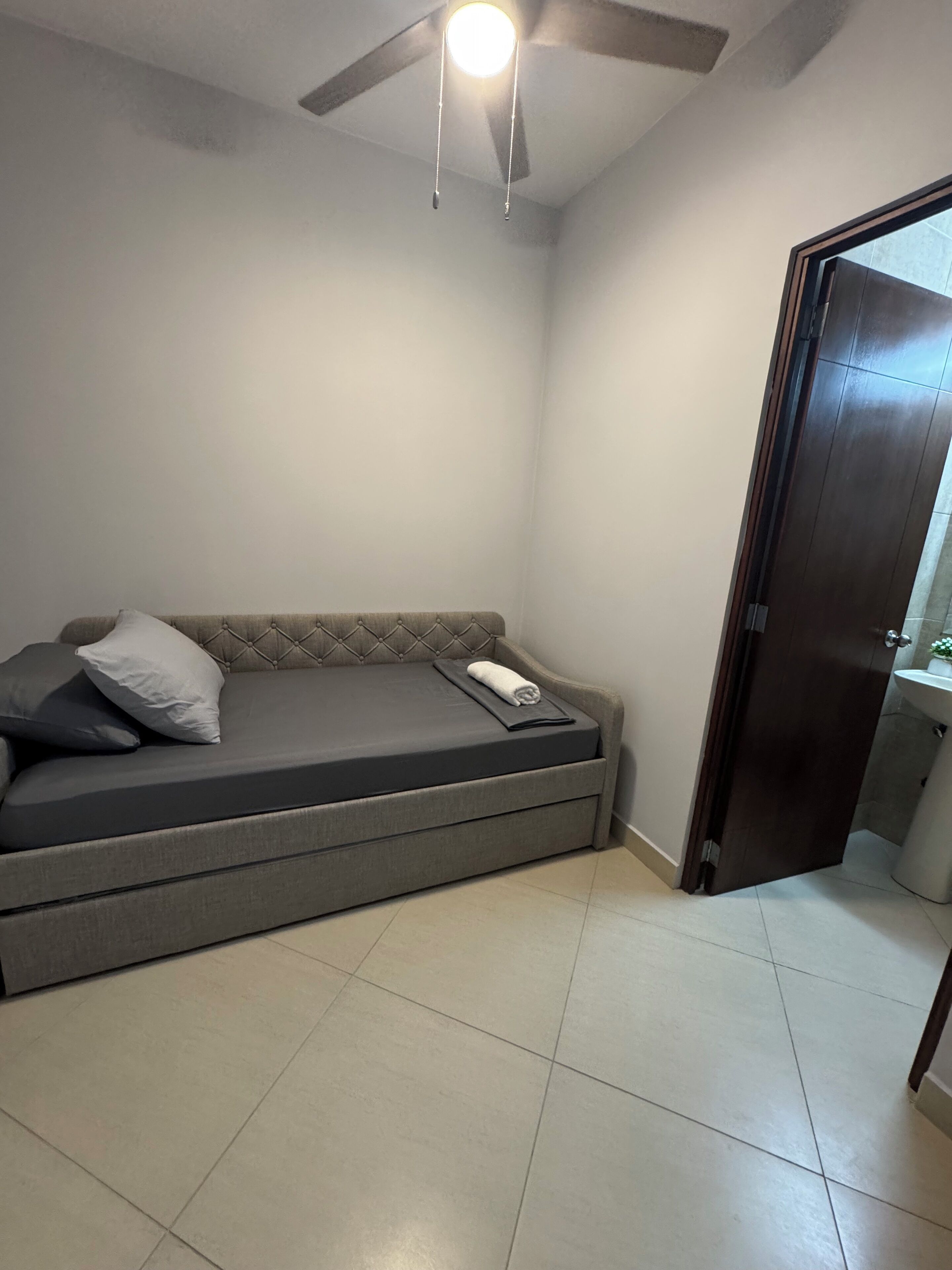 Een kamer met een grijze bank, een plafondventilator en een deur naar een andere ruimte.