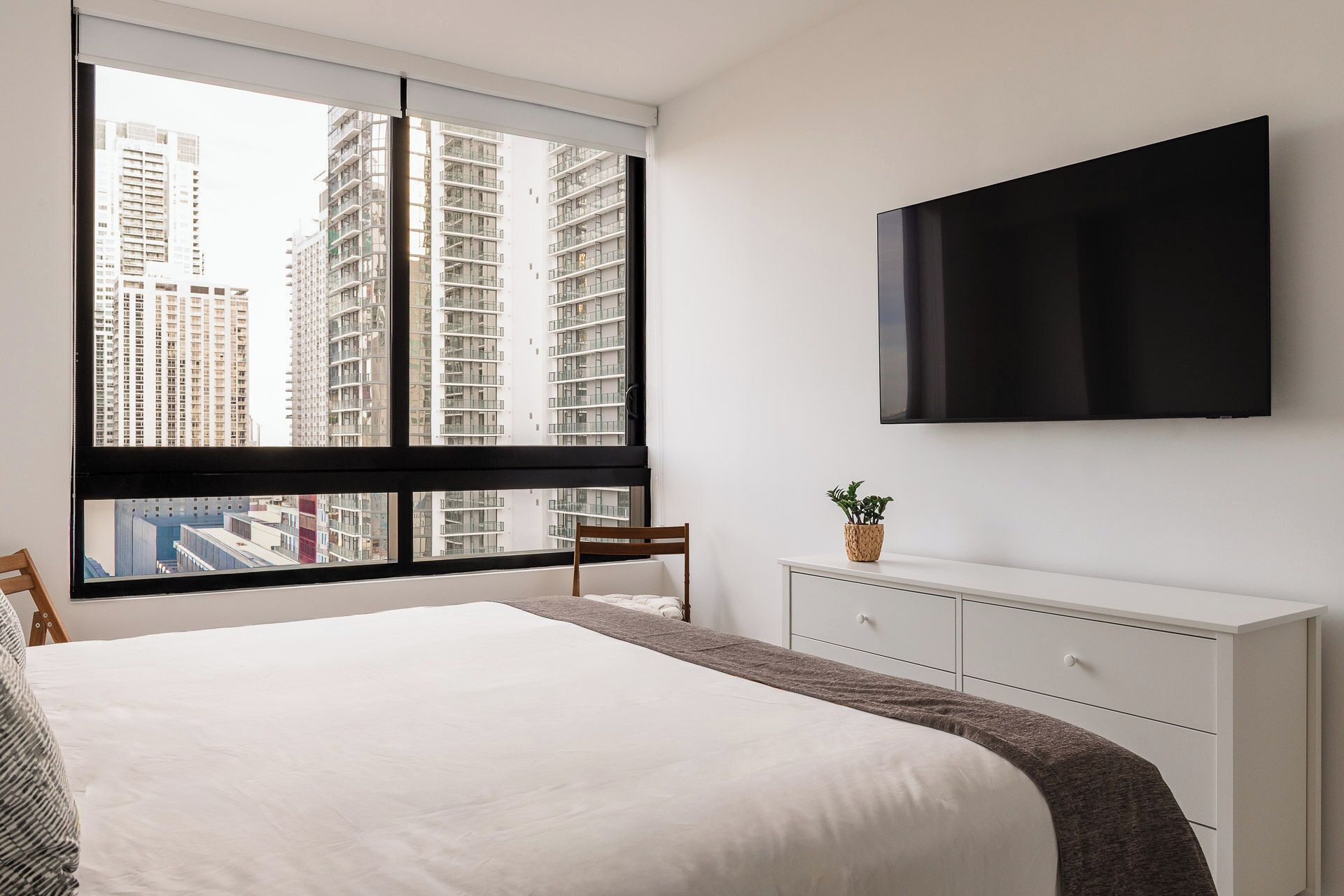Un dormitorio con una cama grande, un televisor montado en la pared, una ventana con vistas a edificios altos y un aparador blanco.