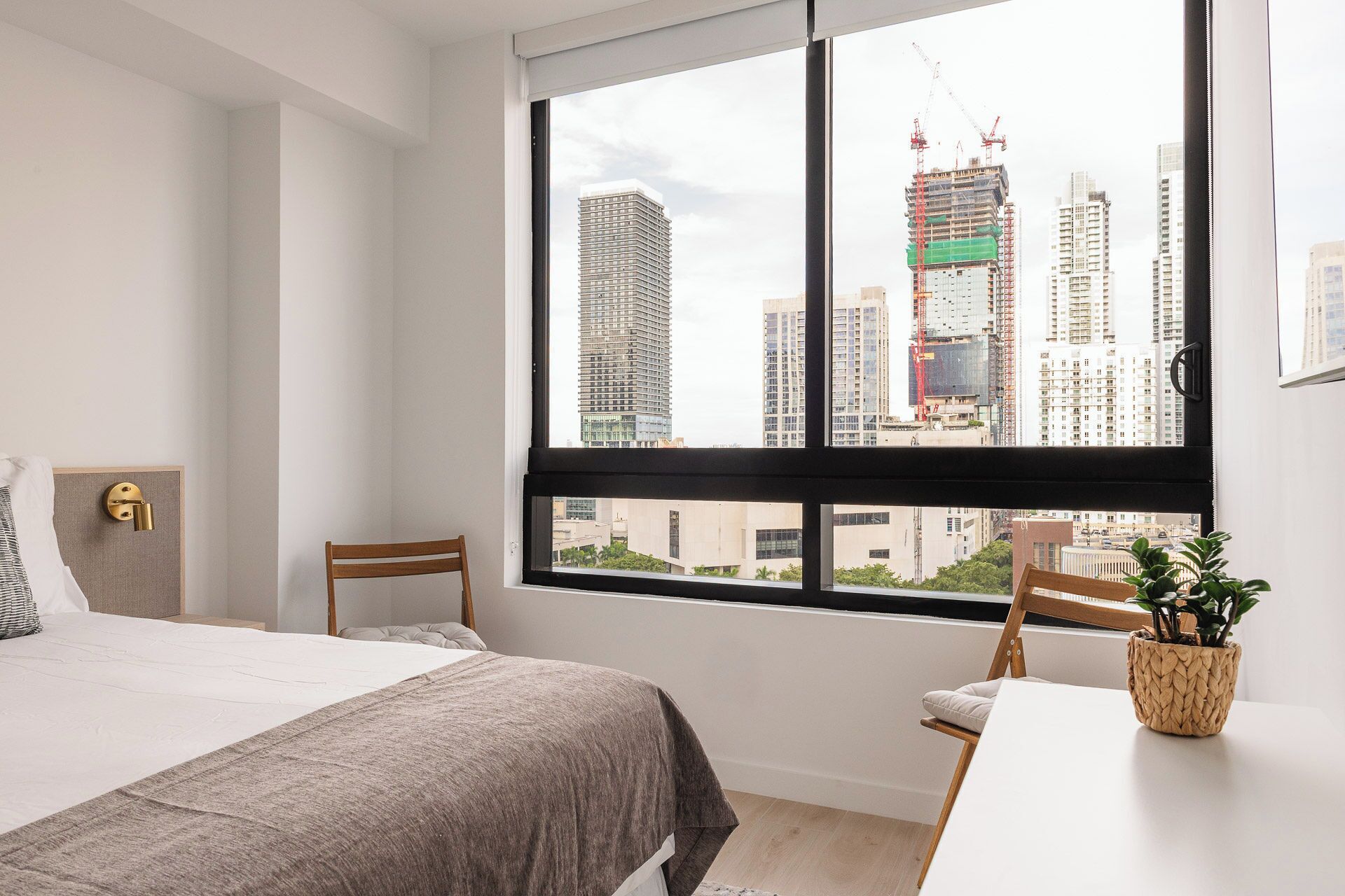 A modern hotel room with a large window, a bed, a chair, a small table, and a potted plant.