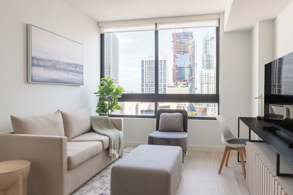 Una sala de estar moderna con un sofá, un reposapiés y un sillón, un televisor y un ventanal con vistas a la ciudad.
