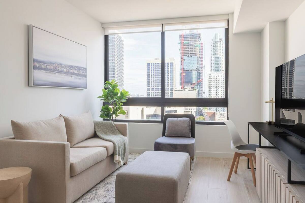 Ein modernes Wohnzimmer mit einer Couch, einem Hocker und einem kleinen Tisch. Ein großes Fenster bietet Blick auf hohe Gebäude.
