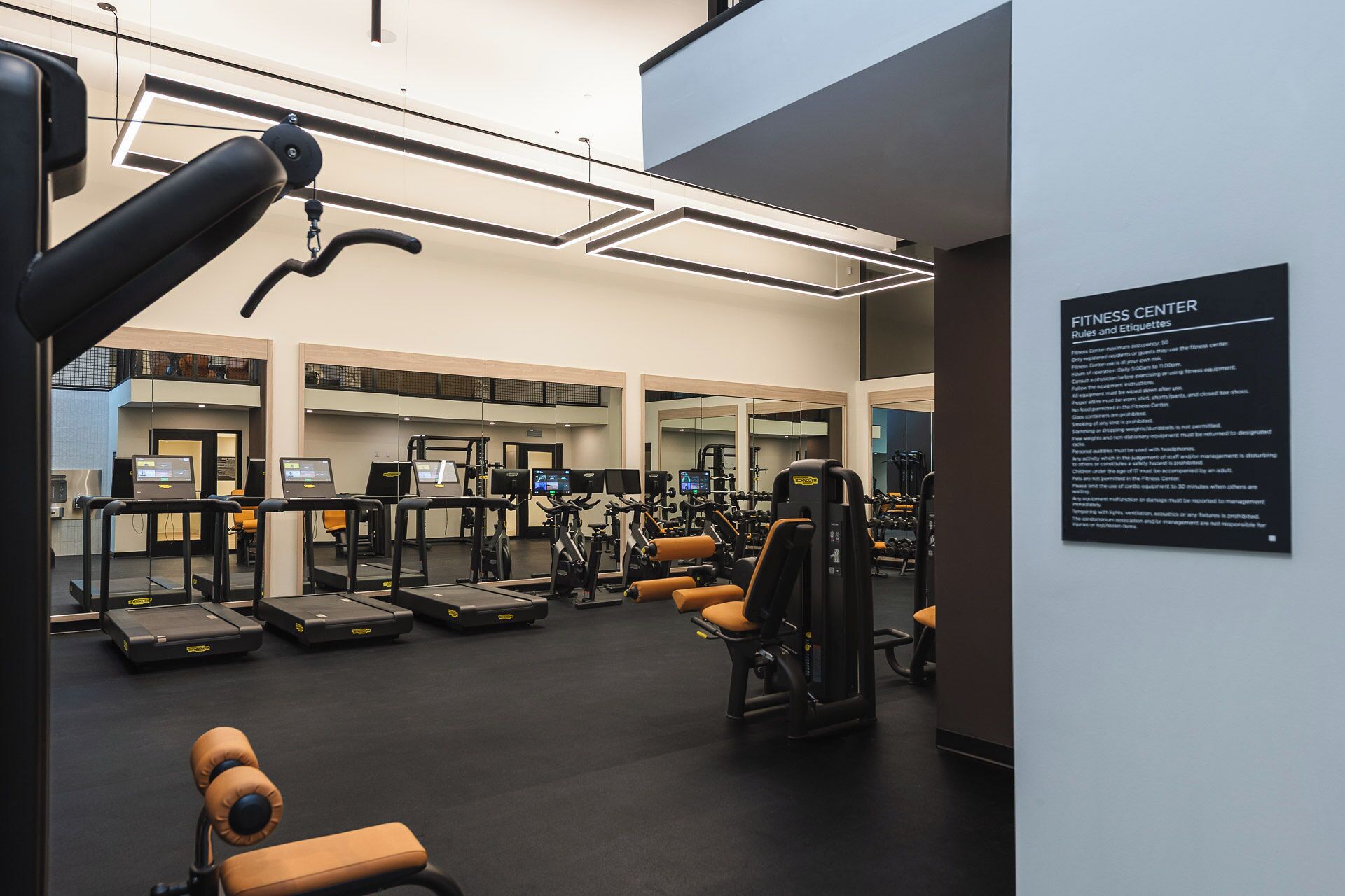 A modern fitness center with various exercise equipment, including treadmills and weight machines, and a sign listing rules and etiquette.