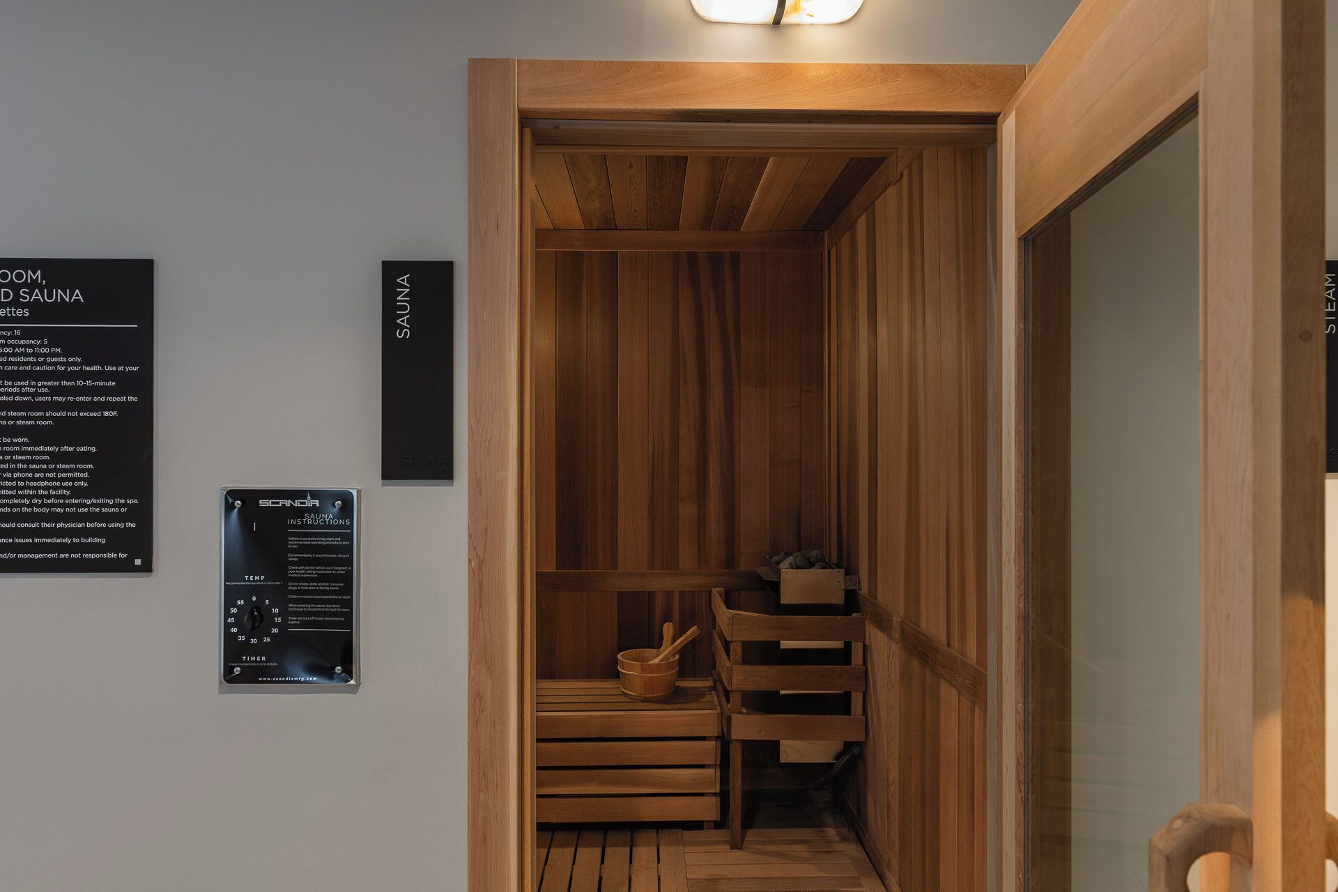 A sauna room with wooden walls and benches, a wooden door, and a light fixture above the door. There are signs on the wall providing instructions and information about the sauna.