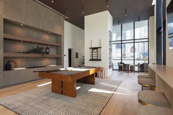 A modern indoor pool table with a wooden finish, surrounded by a patterned rug. There are shelves with decorative items and a large window providing a view of the cityscape. - District 225 by Nomada (Miami)