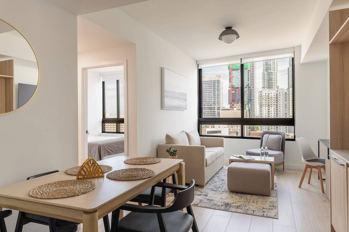 Ein modernes Apartment mit Essbereich, Wohnzimmer und Küche. Es verfügt über große Fenster mit Blick auf die Stadt, einen runden Spiegel und Hochhäuser im Hintergrund.