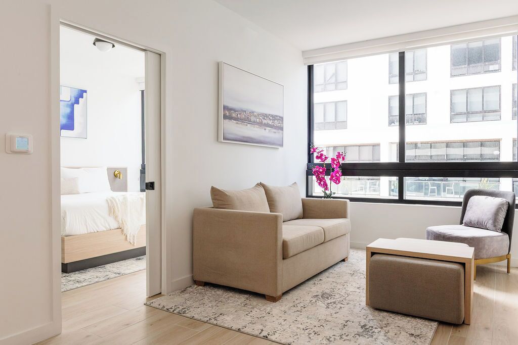 A modern living room with a sofa, ottoman, and a chair. There is a large window with a view of buildings.