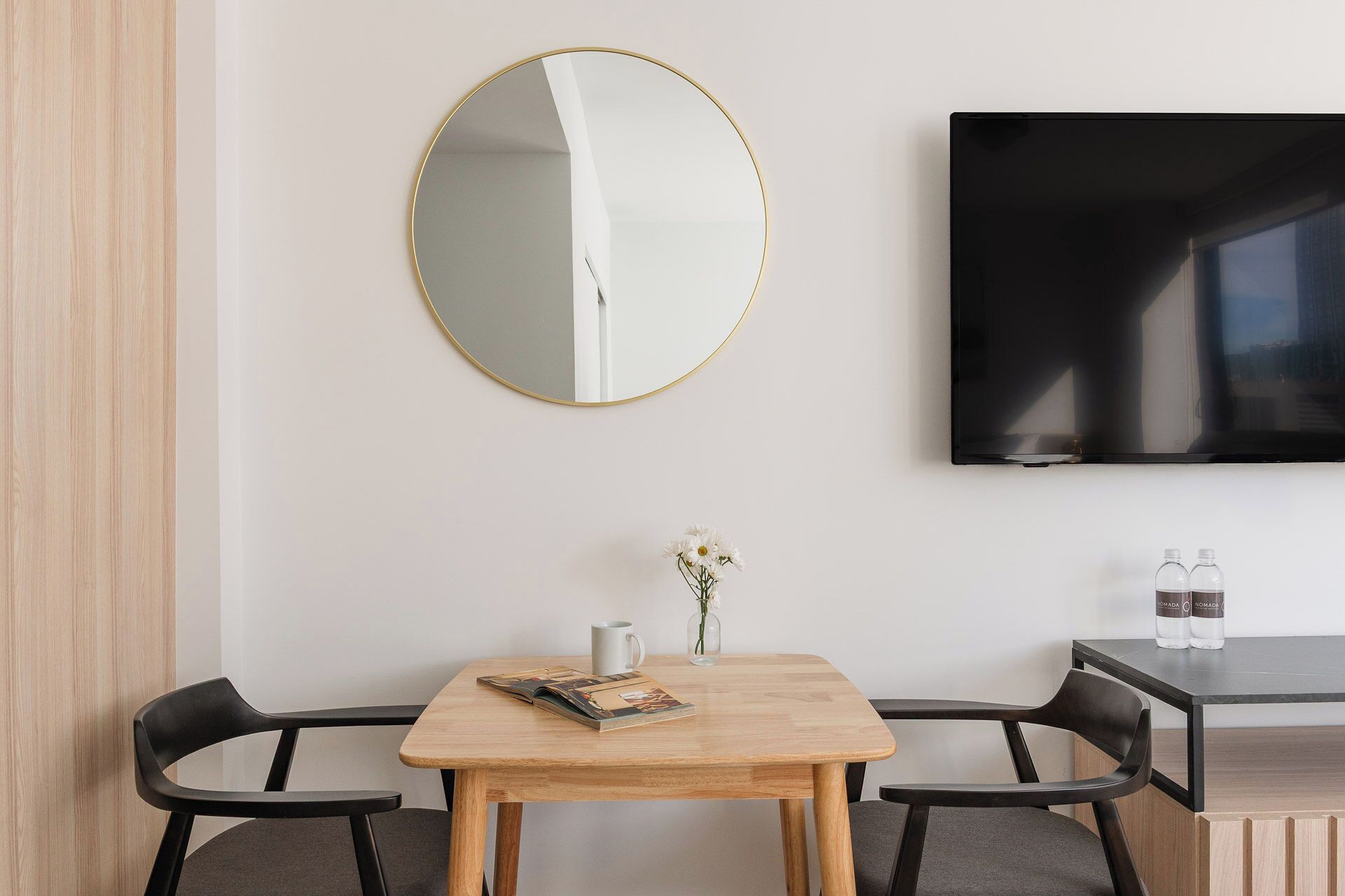 A modern room with a wooden table, two black chairs, a round mirror, a flat-screen TV, and a small vase with flowers.