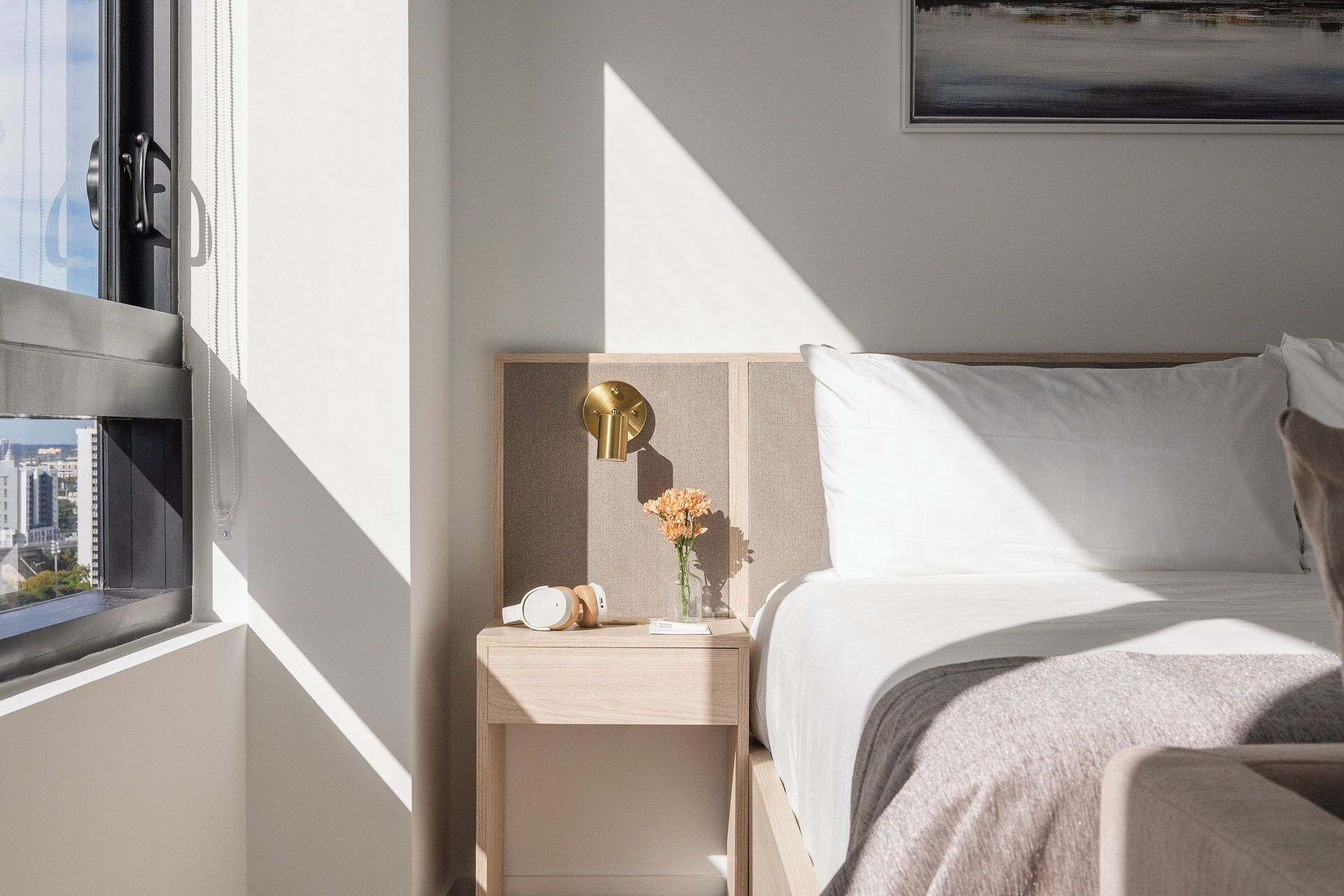 Una habitación de hotel moderna con una cama, una mesita de noche con una lámpara y un ventanal con vistas a edificios.