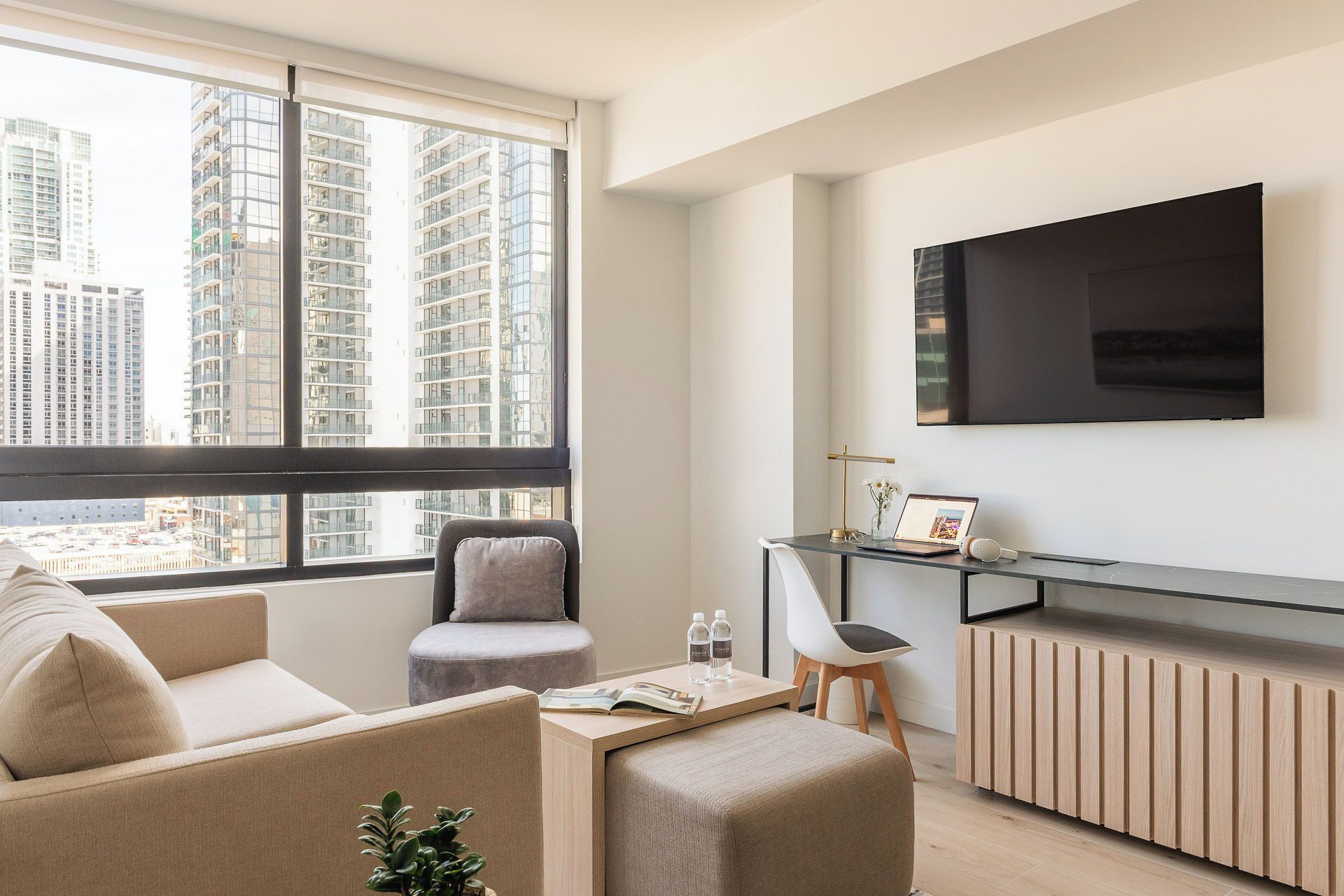 A modern living room with a sofa, a small table with a laptop, a chair, and a large window with a city view.