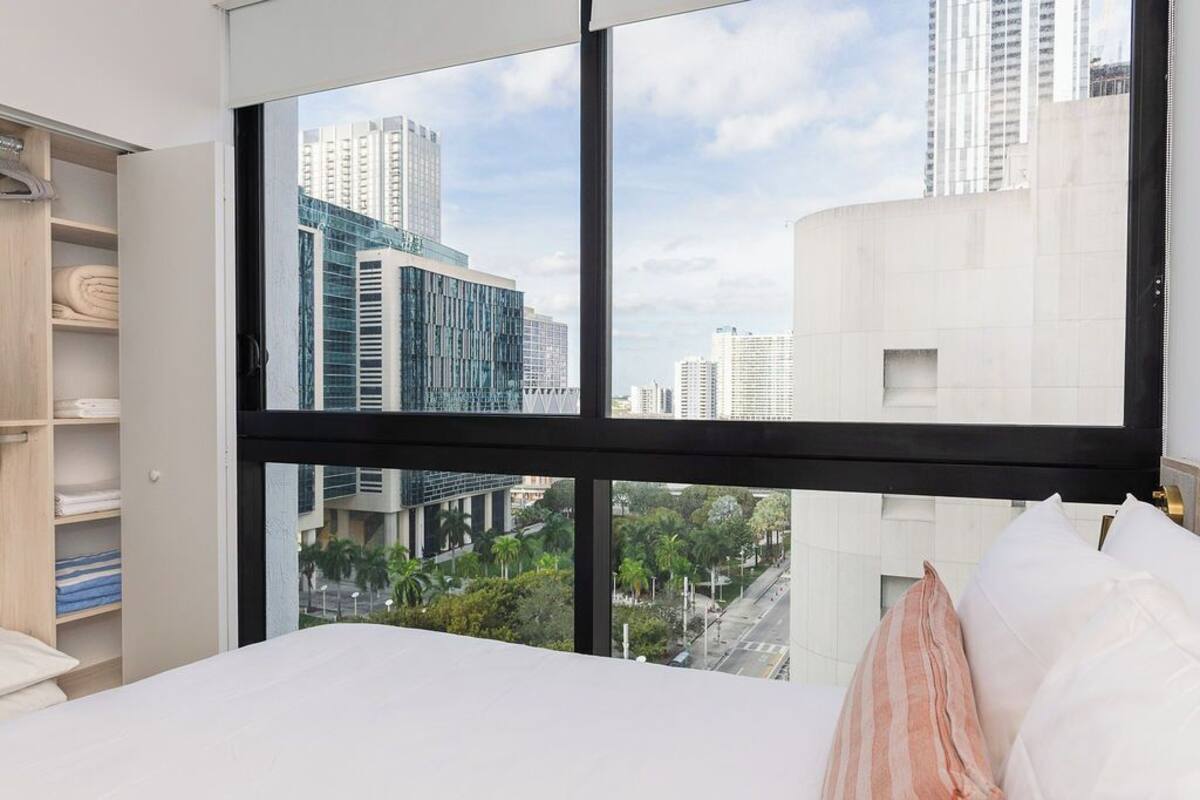 Ein Schlafzimmer mit großem Fenster und Blick auf die Stadt, einem Bett mit weißer Bettwäsche und einem hölzernen Kleiderschrank.
