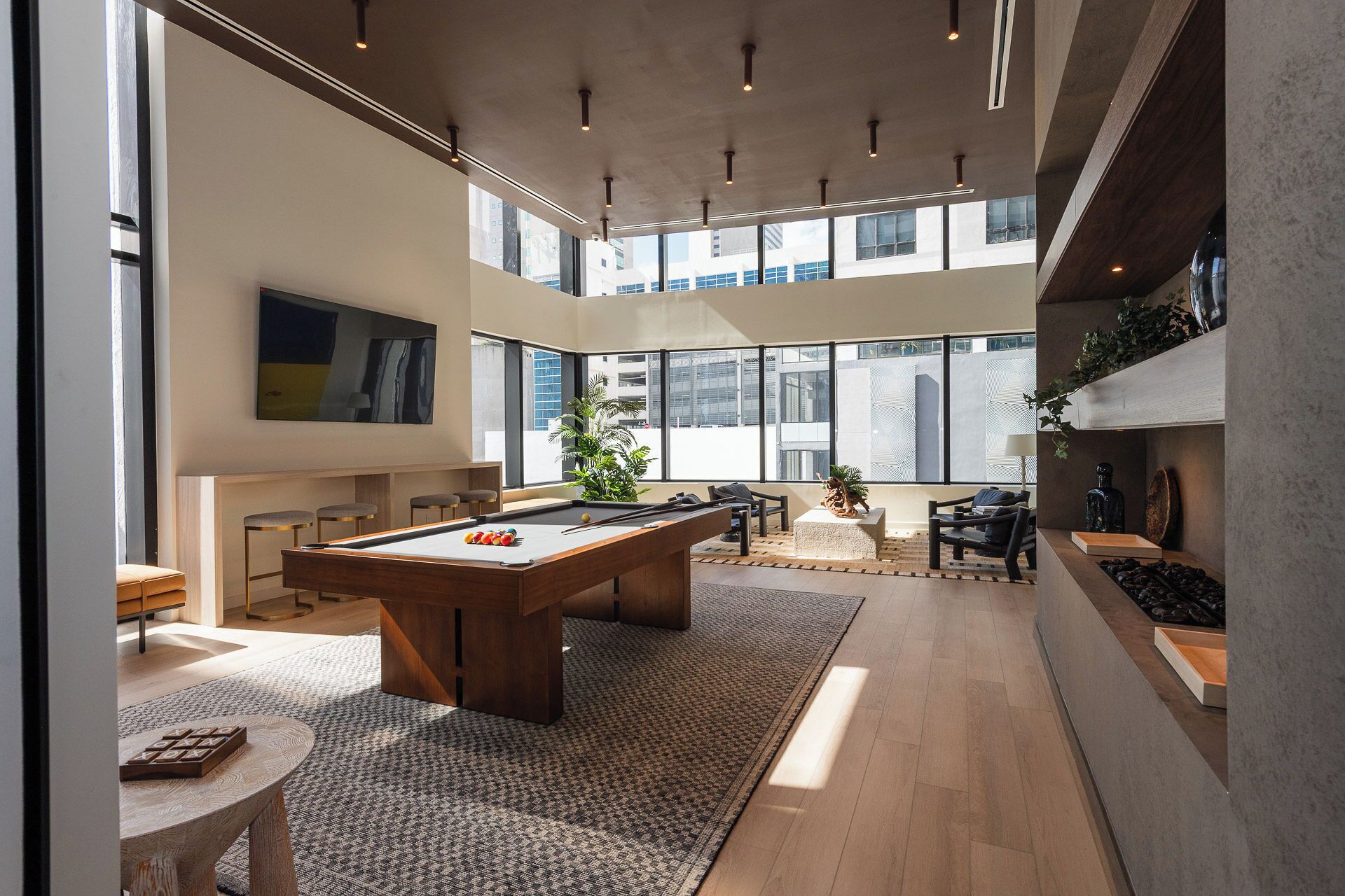 A modern indoor space with a pool table, seating area, and large windows offering views of the city.