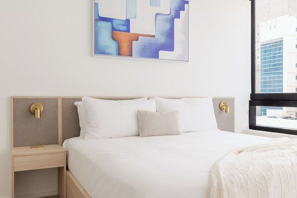Moderni hotellihuone, jossa on suuri sänky, yöpöytä, seinälle ripustettu taideteos ja ikkuna, josta avautuu näkymä kaupunkiin.