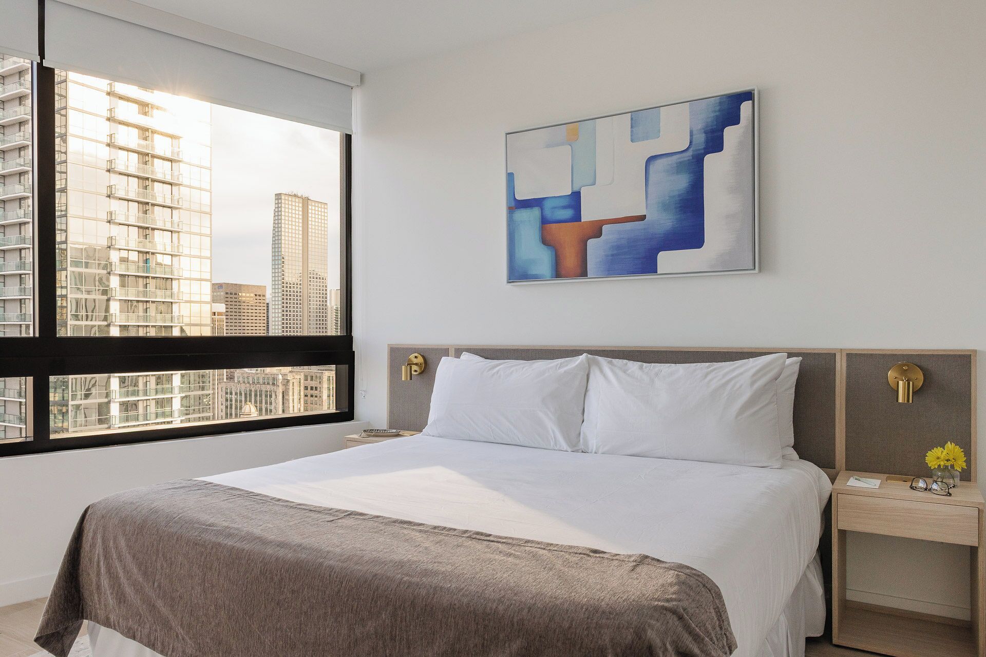 A modern hotel room with a large bed, a city view, and a contemporary abstract painting.