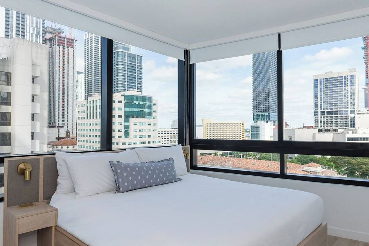 Ein modernes Hotelzimmer mit einem großen Bett, einem Nachttisch und Blick auf die Skyline der Stadt durch große Fenster.