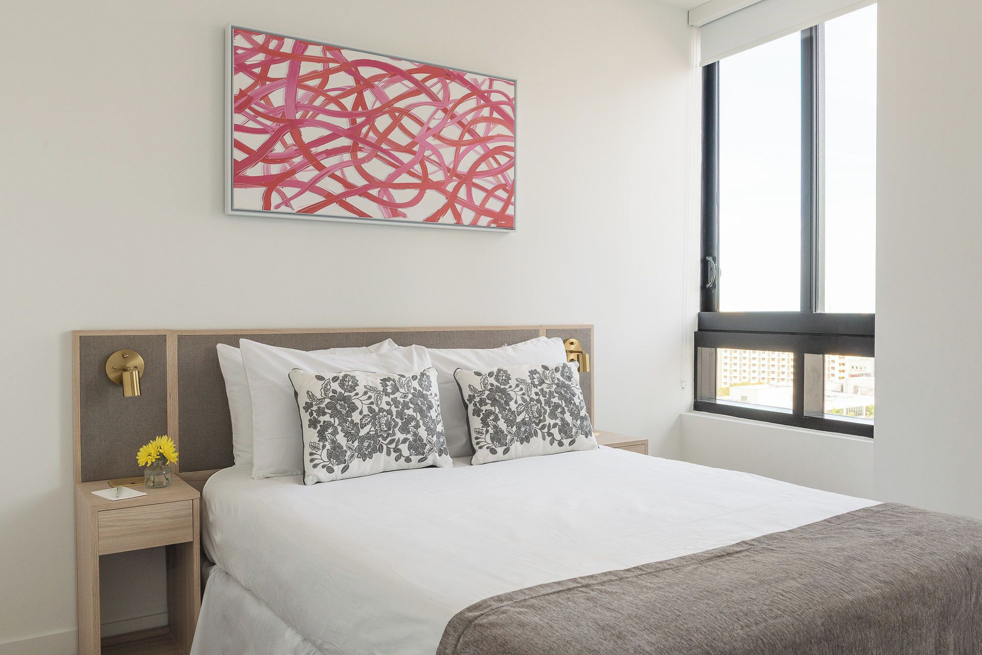 A hotel room with a large bed, two nightstands, a window with blinds, and a red abstract painting on the wall.