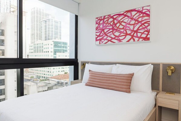 A hotel room with a large window, a bed with white and pink bedding, a modern abstract painting, and a nightstand with a lamp. - District 225 by Nomada (Miami)