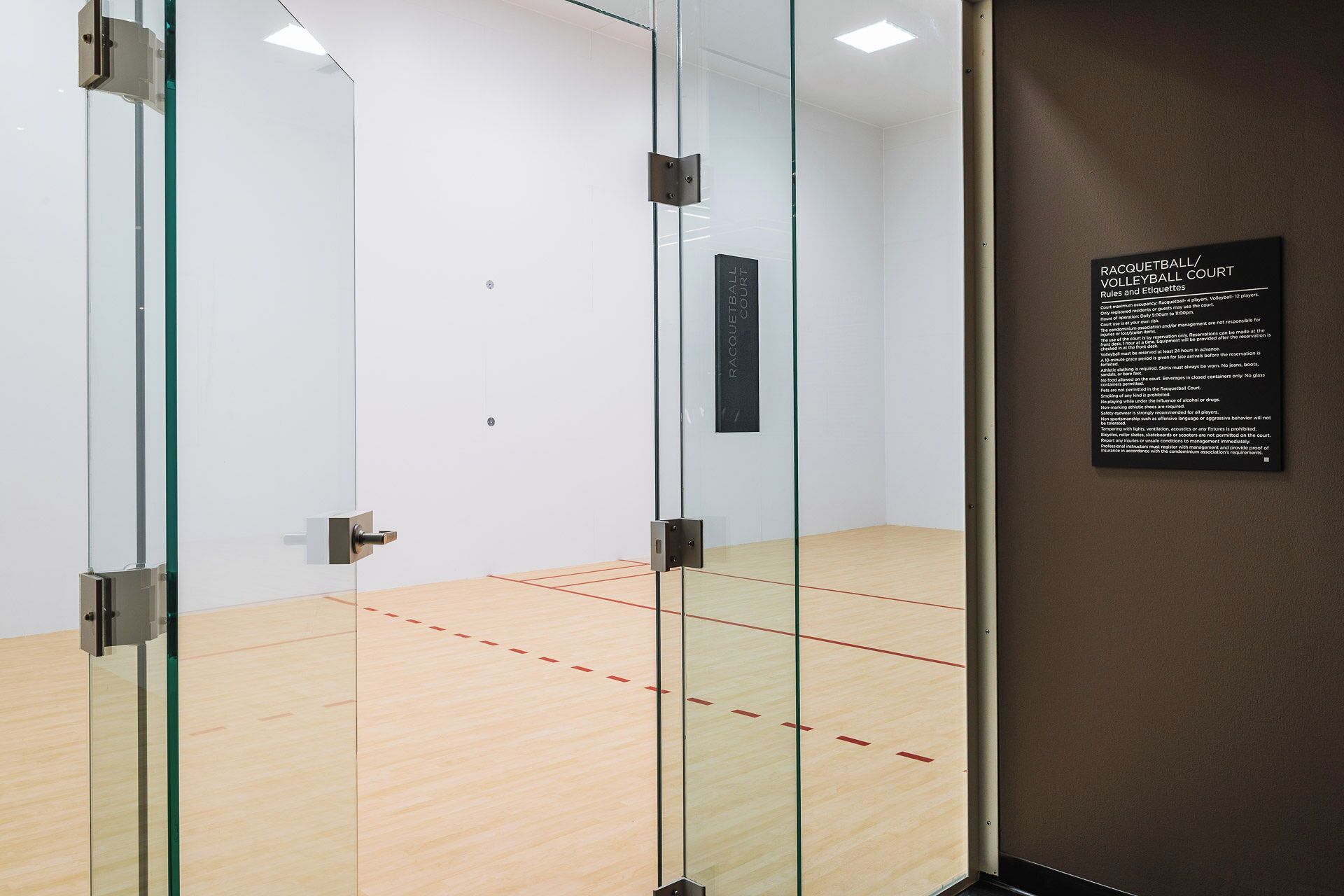 A squash court with glass walls, a wooden floor, and a sign detailing court rules and etiquette.