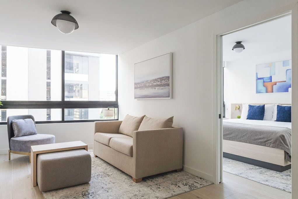 A modern living room with a sofa, ottoman, and armchair, a large window with a view of buildings, and a door leading to a bedroom with a bed and artwork.