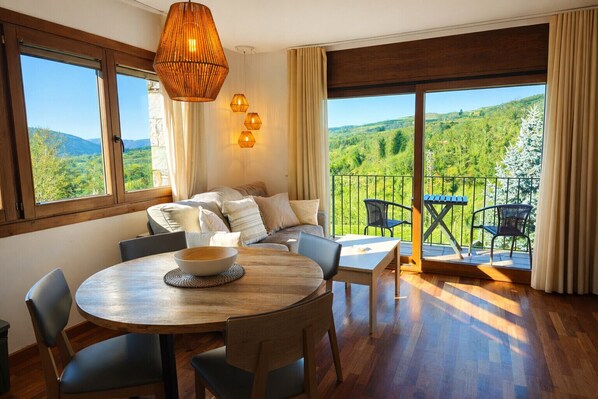 En moderne stue med sofa, spisebord, stole og en balkon med udsigt til grønne områder, samt et stort vindue.