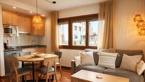A modern kitchen with wooden cabinets, a dining table with chairs, a grey sofa with cushions, and a window with curtains. - Vall de Muntanyes - Costabona (Molló)