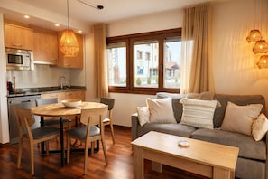 A modern kitchen with wooden cabinets, a dining table with chairs, a grey sofa with cushions, and a window with curtains. - Vall de Muntanyes - Costabona (Molló)