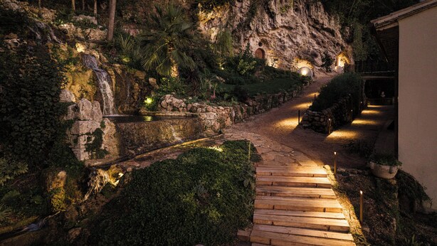 Ein Steinweg führt zu einem rustikalen Gebäude mit einem Wasserfall und beleuchteten Lichtern.