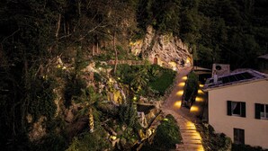Ein Bergpfad führt zu einem rustikalen Gebäude mit Natursteinfassade, umgeben von dichtem Wald und beleuchtet durch warmes Licht.