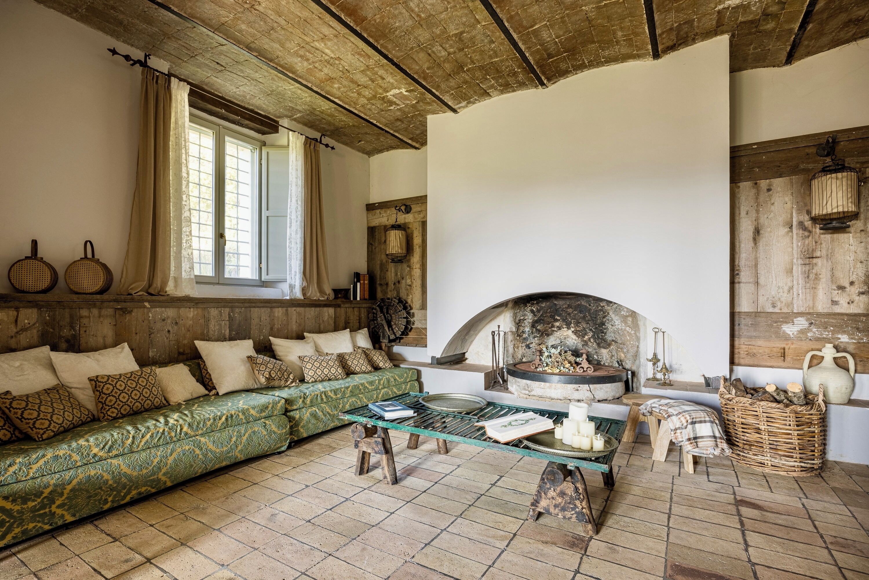 A rustic living room with a green patterned sofa, a wooden coffee table, a fireplace, and a window with curtains.