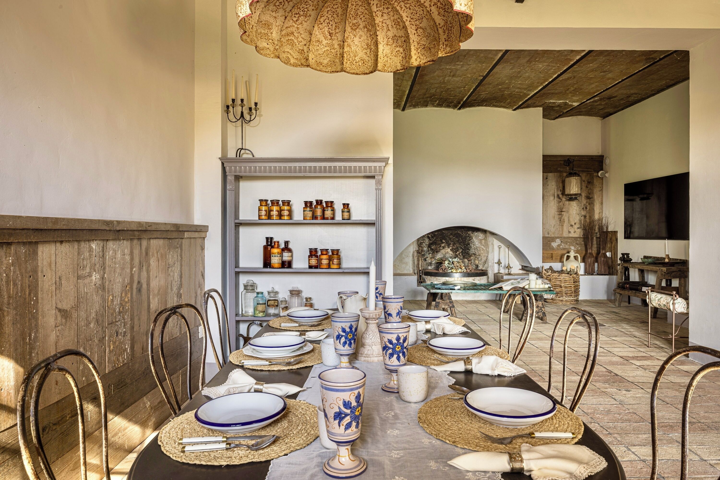 A dining area with a long table set for six, surrounded by metal chairs. A wooden wall and a fireplace are visible in the background.