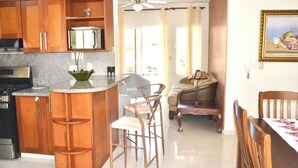 Una cocina con gabinetes de madera, microondas empotrado y estufa a gas. Hay dos taburetes de bar y una mesa de comedor con sillas.