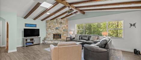A spacious living room with a stone fireplace, a flat-screen TV, and a large window with blinds.