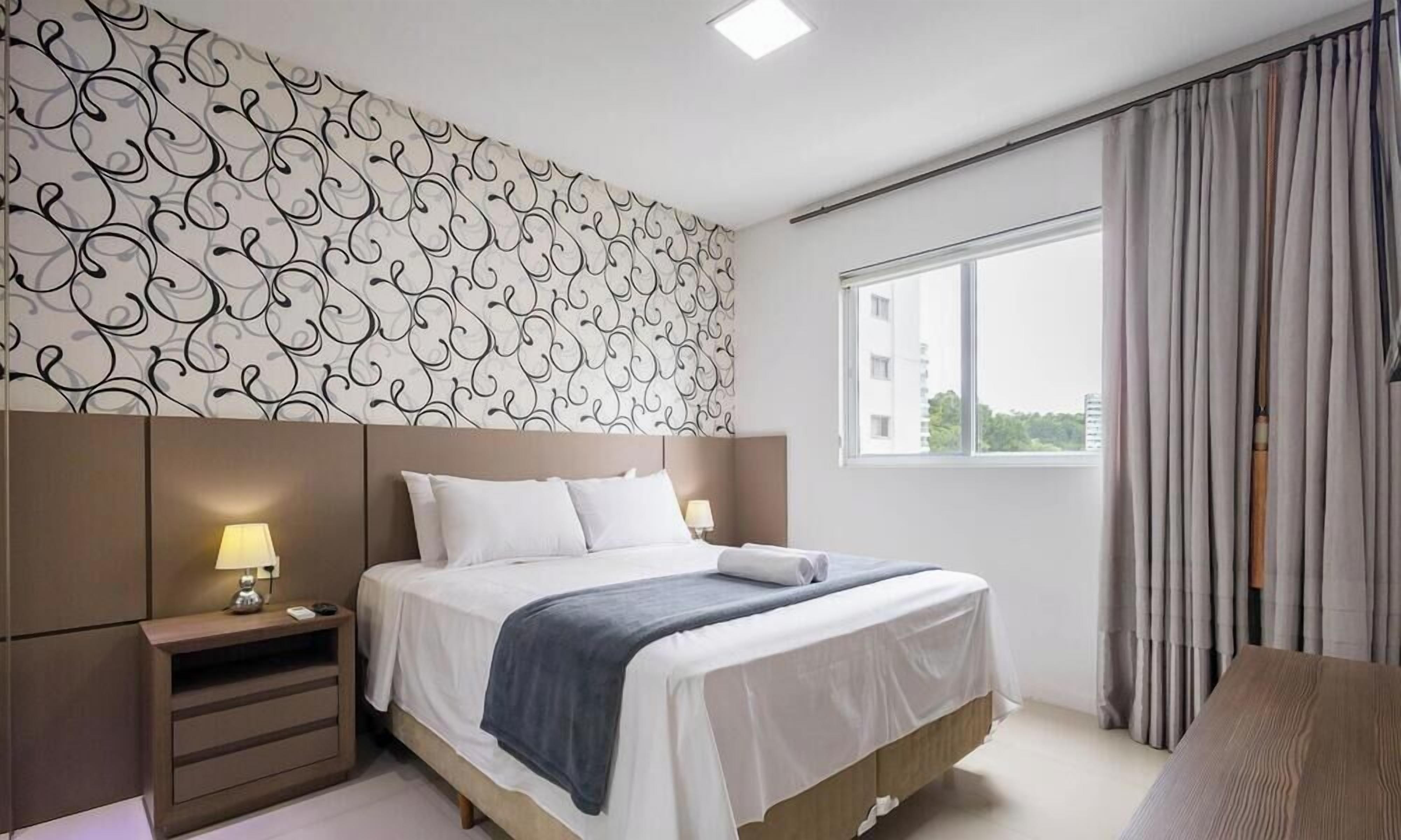 A hotel room with a bed, bedside table, lamp, and a window with curtains.