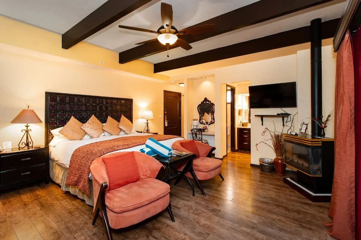 A hotel room with a bed, two chairs, a desk, a ceiling fan, a TV, and a fireplace.