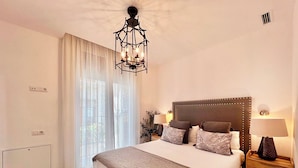 A bedroom with a large bed, a chandelier, a bedside table with a lamp, and a window with curtains. - Apartamentos Realejo Campo Príncipe 3000 (Granada)