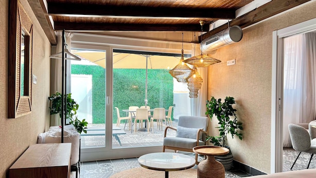 A hotel room with a large bed, a round coffee table, and a seating area with chairs and a table. There is a large window with a view of a green hedge outside. - Apartamentos Realejo Campo Príncipe 3000 (Granada)