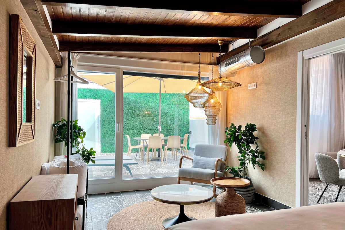 Ein Hotelzimmer mit einem großen Bett, einem runden Couchtisch und einer Sitzecke mit Stühlen und einem Tisch. Ein großes Fenster bietet Blick auf einen grünen Heckenstreifen.