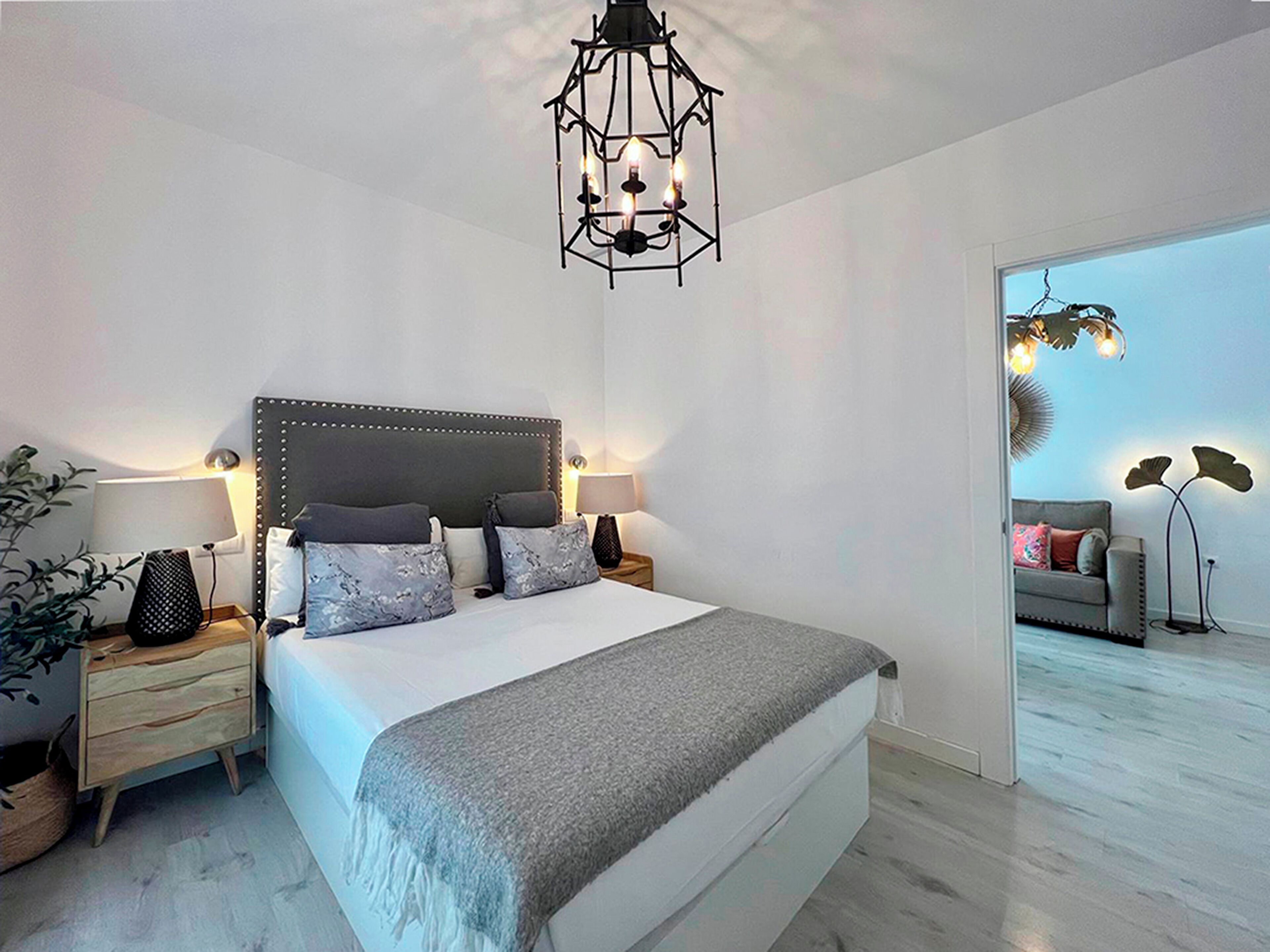 Et moderne soveværelse med en stor seng, sengeborde, en hængende lampe og udsigt til et opholdsområde gennem en åben dør.