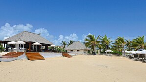 A beachfront resort with thatched-roof buildings, outdoor seating, and palm trees. - Spacious and Elegant 2BR Condo, King size Bed, Full AC, Pool, Steps to Hard Rock (Punta Cana)