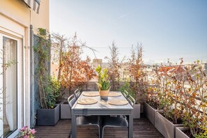Eine Dachterrasse mit Tisch für zwei Personen, umgeben von Topfpflanzen und Blick auf eine Stadtlandschaft.