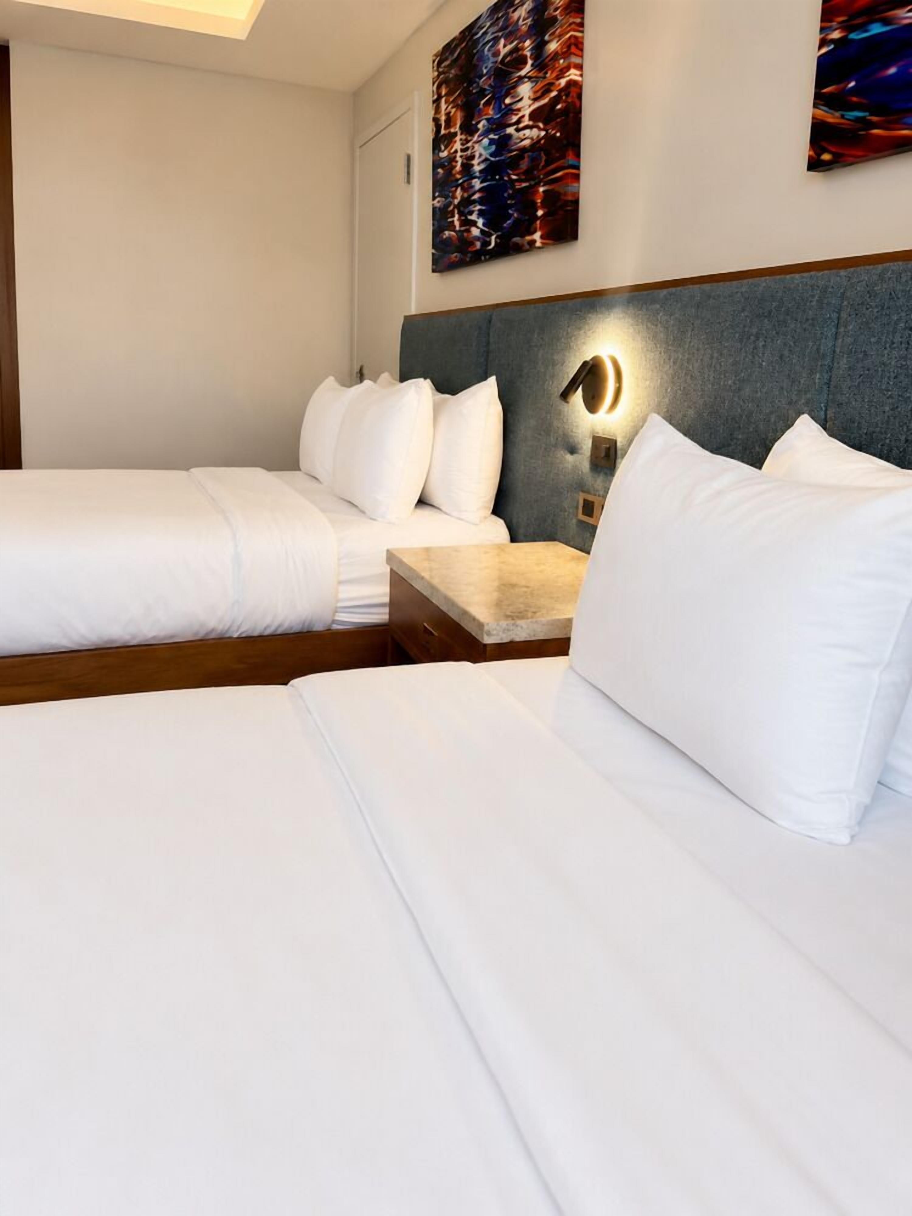 A hotel room with two beds, a nightstand, and a wall-mounted lamp.