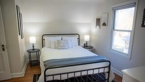 Ein Schlafzimmer mit einem Metallbettgestell, weißer Bettwäsche, zwei Nachttischlampen, einem Fenster und einer Tür.