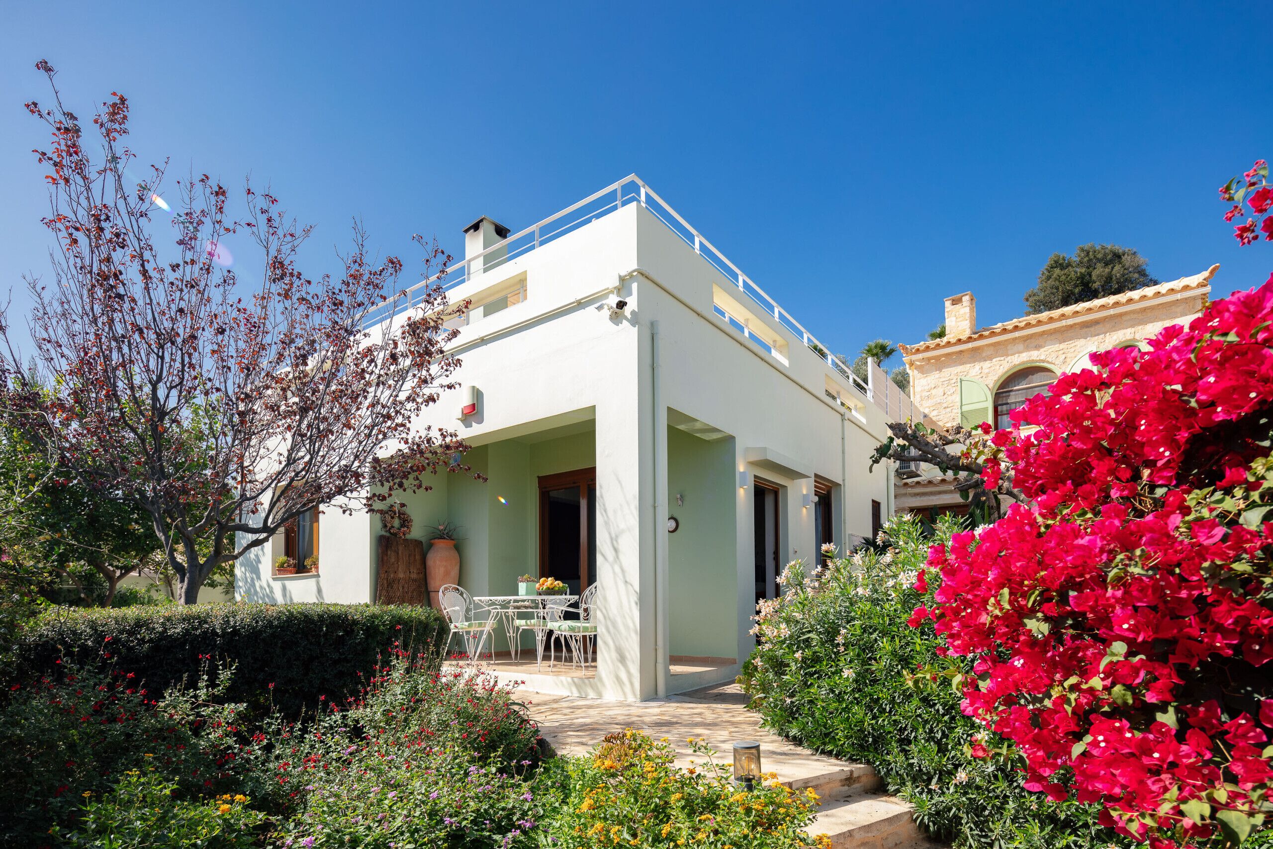 A white building with a balcony, surrounded by a garden with various plants and flowers.