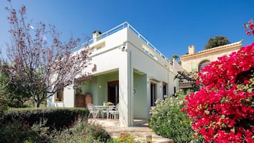 A white building with a balcony, surrounded by a garden with various plants and flowers.