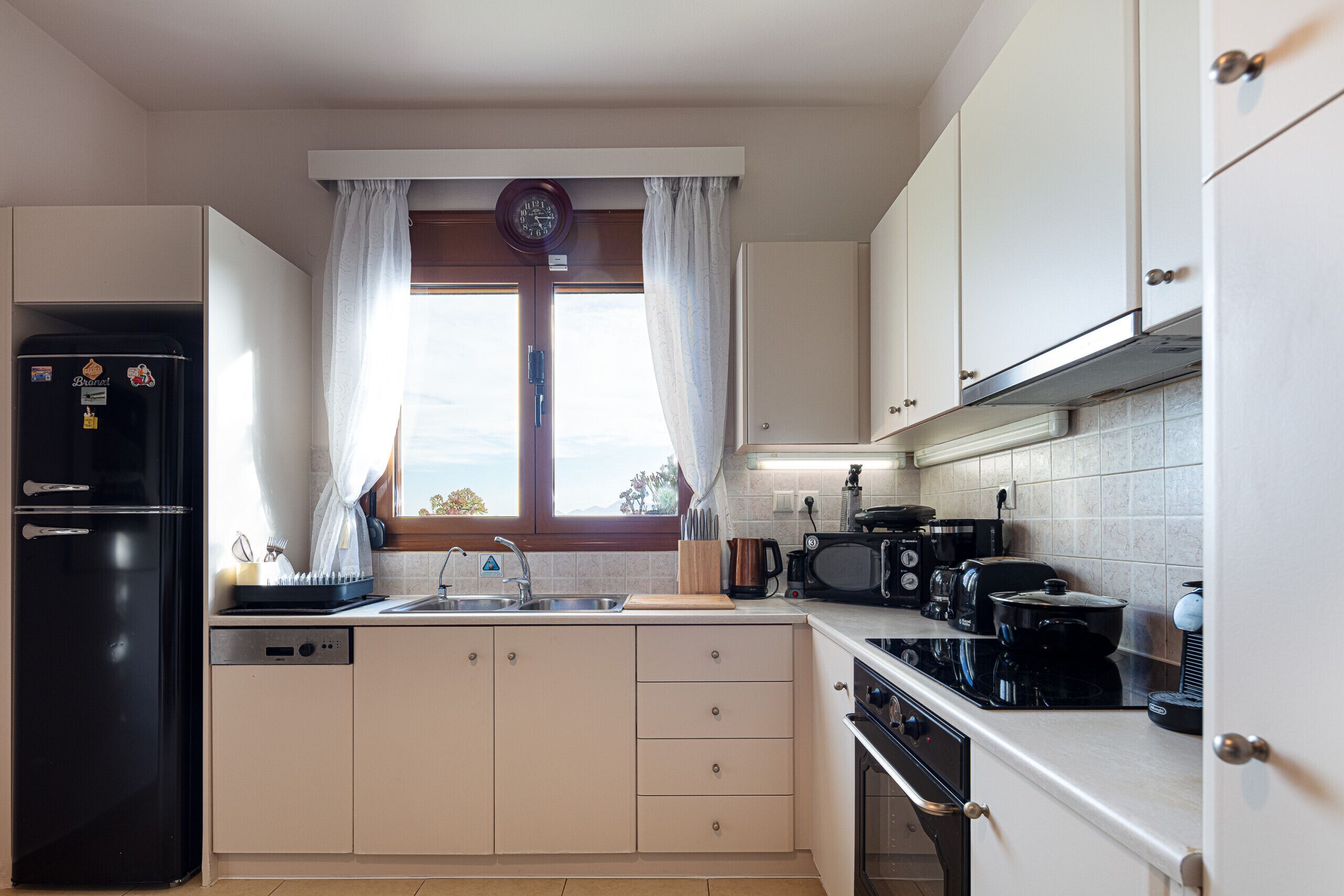 Uma cozinha moderna com geladeira preta, micro-ondas, fogão e lava-louças. Há uma janela com cortinas e um relógio na parede.